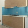 Cucina con mobili in legno chiaro, piastrelle blu e un piccolo frigorifero bianco.