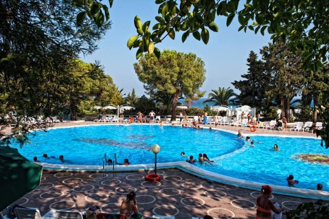 Piscina esterna con molti bagnanti e alberi intorno.