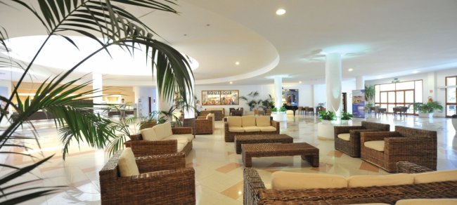 Area lounge con divani in rattan e cuscini chiari, piante sparse.