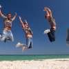 Sei persone saltano sulla spiaggia sotto un cielo blu.